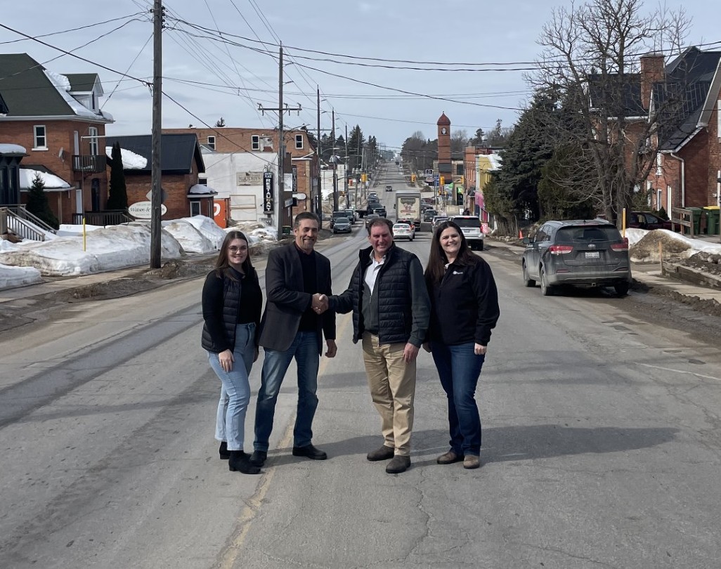 Four people stand on a town street, two men in the center shaking hands, conveying a sense of agreement. Snow-lined sidewalks, traffic, and buildings surround them.